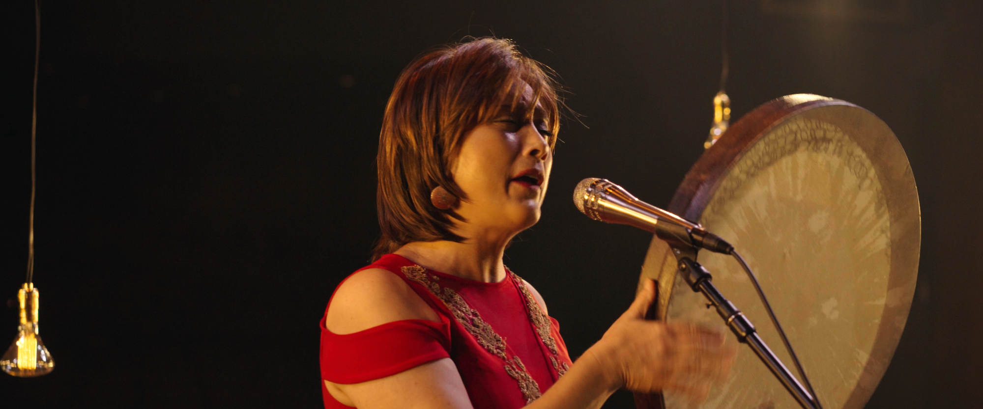 A woman in a red dress passionately sings into a microphone while playing a large frame drum. Hanging bulbs illuminate the stage, adding a warm glow to the scene.