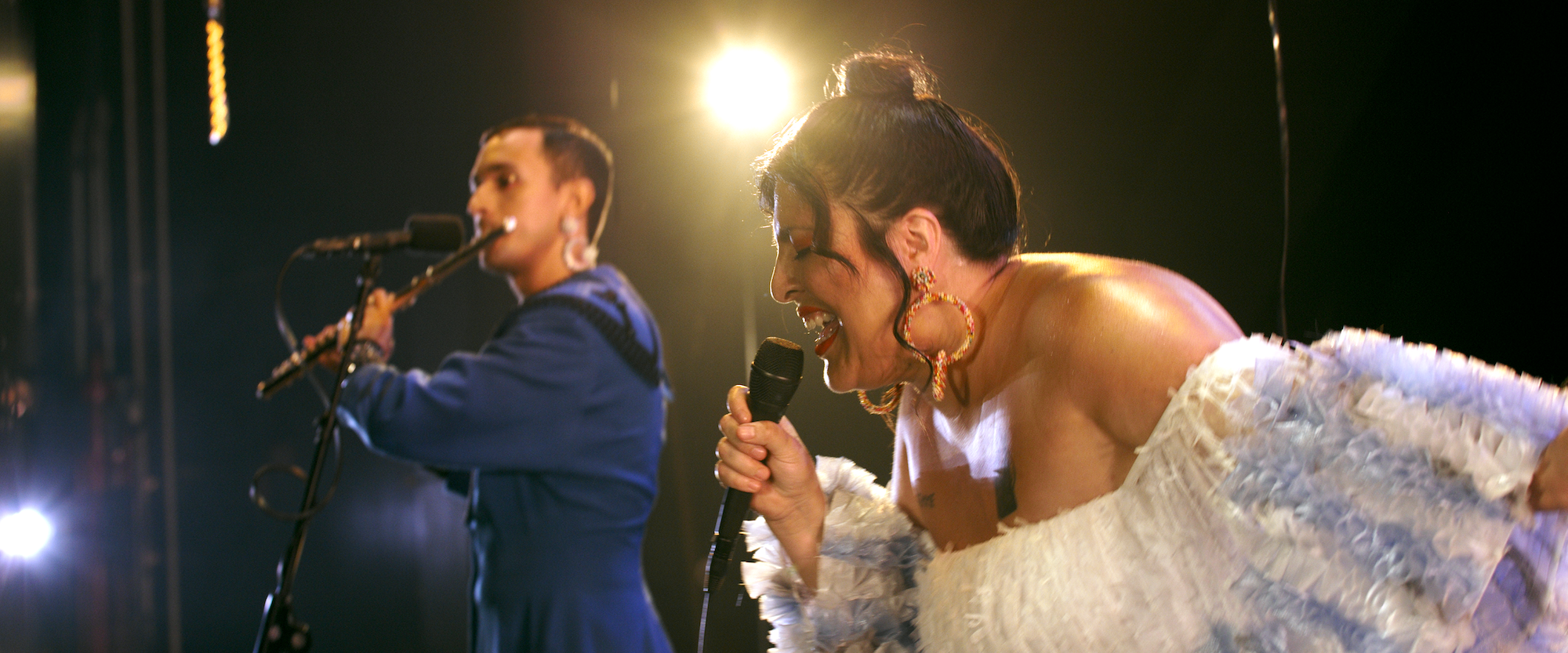 Two performers on stage sing into microphones. One in a blue suit stands to the left, while the other in a white, feathered outfit with large hoop earrings sings passionately in the foreground. Bright stage lights shine behind them.