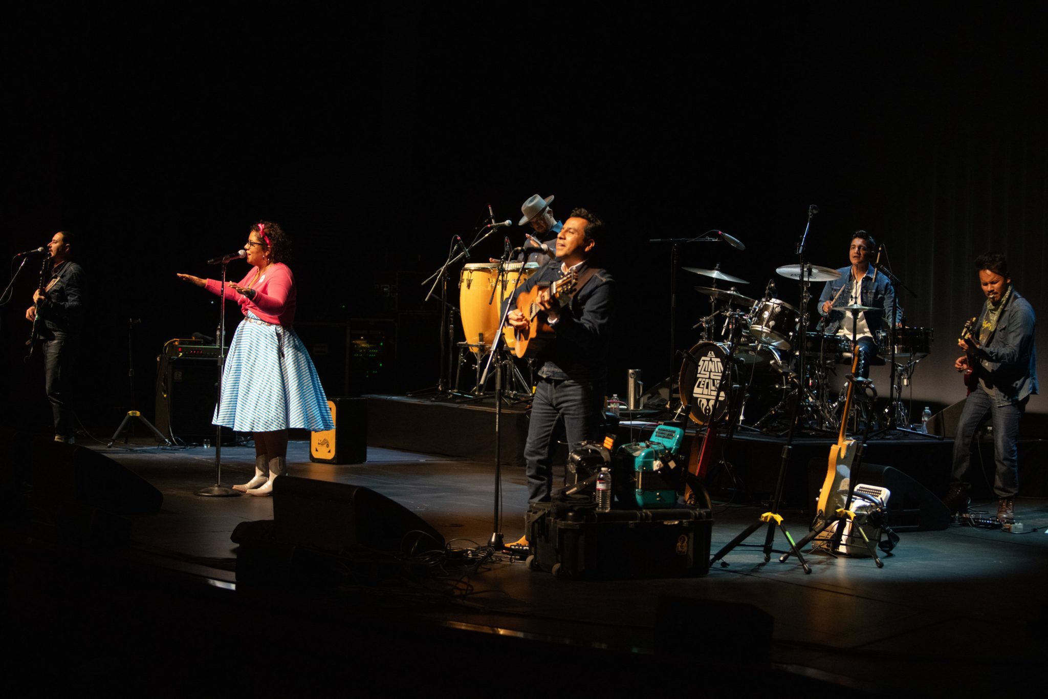 A soulful performance in progress on stage, featuring vocalist La Marisoul with a microphone at the forefront, accompanied by musicians on guitar, percussion, and keyboards.