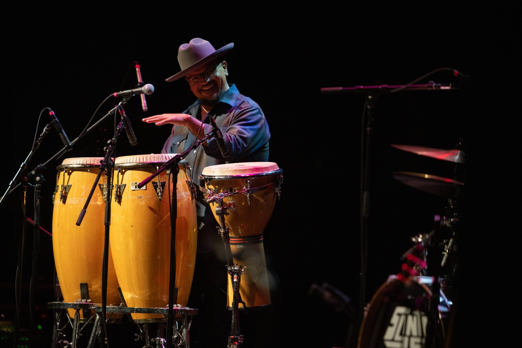 A percussionist in a hat flashing a smile while energetically playing a set of conga drums on stage under moody lighting.