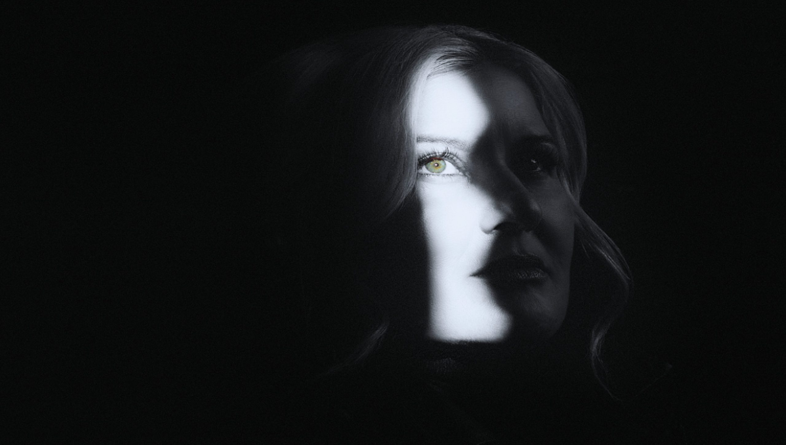 A mysterious portrait of American singer Paula Cole illuminated by a sliver of light, highlighting her intense gaze and delicate facial features amidst the shadows.