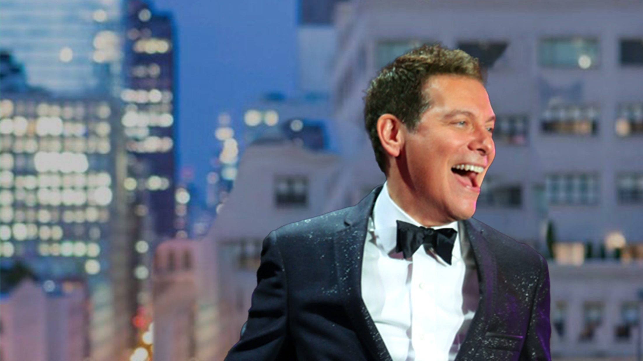 Michael Feinstein in a black tuxedo and bow tie laughing joyously, holding a microphone with a vibrant cityscape at night in the background.