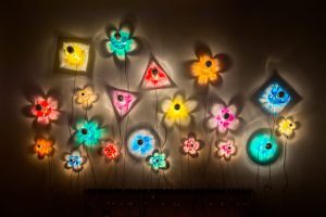 Colorful flower-shaped wall lights in various colors creating a vibrant and whimsical display against a dark background with soft shadows.