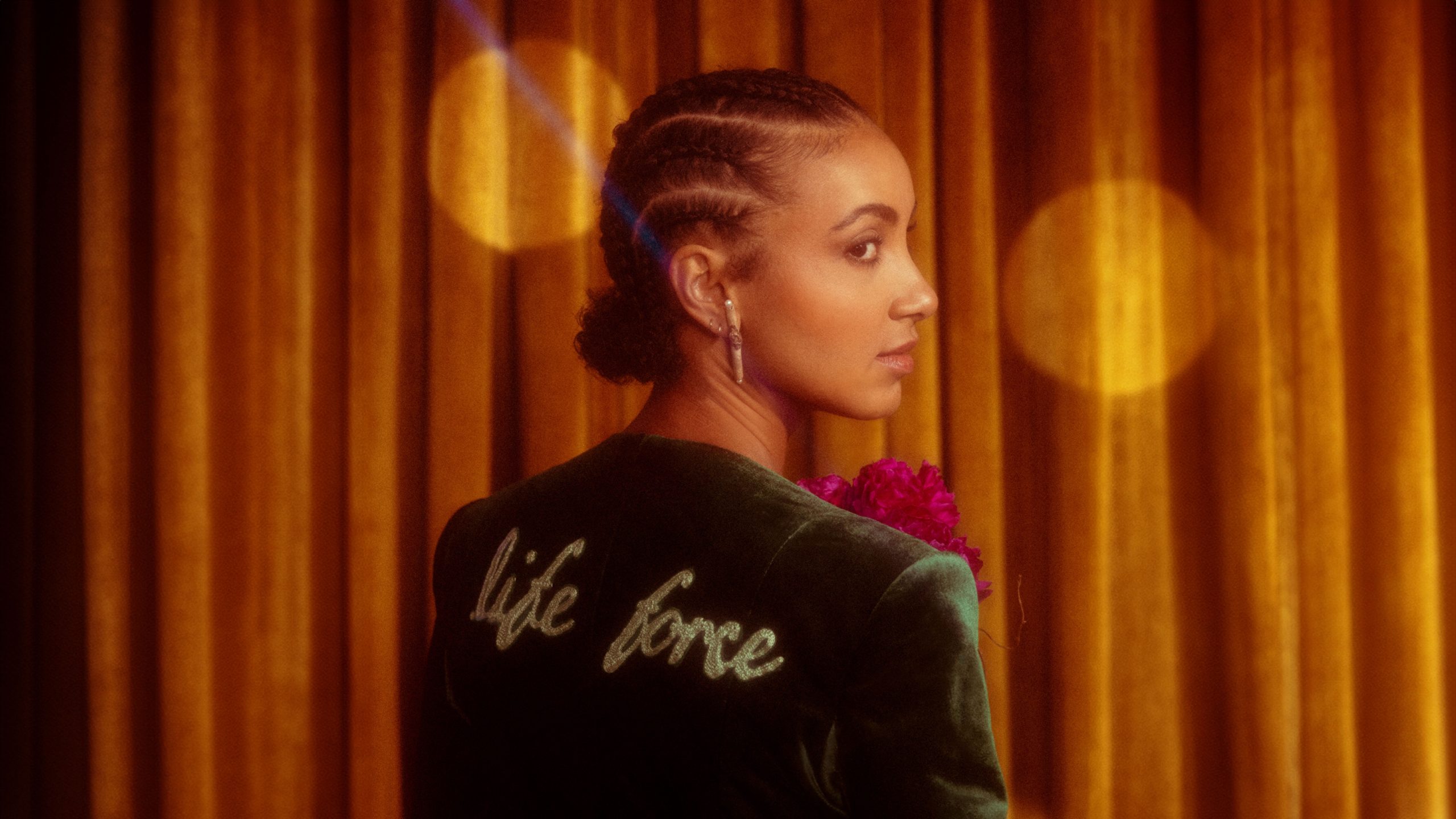 Esperanza Spalding with braided hair and a flower gazes thoughtfully to the side against a backdrop of warm golden drapery, illuminated by a soft, diffused light.