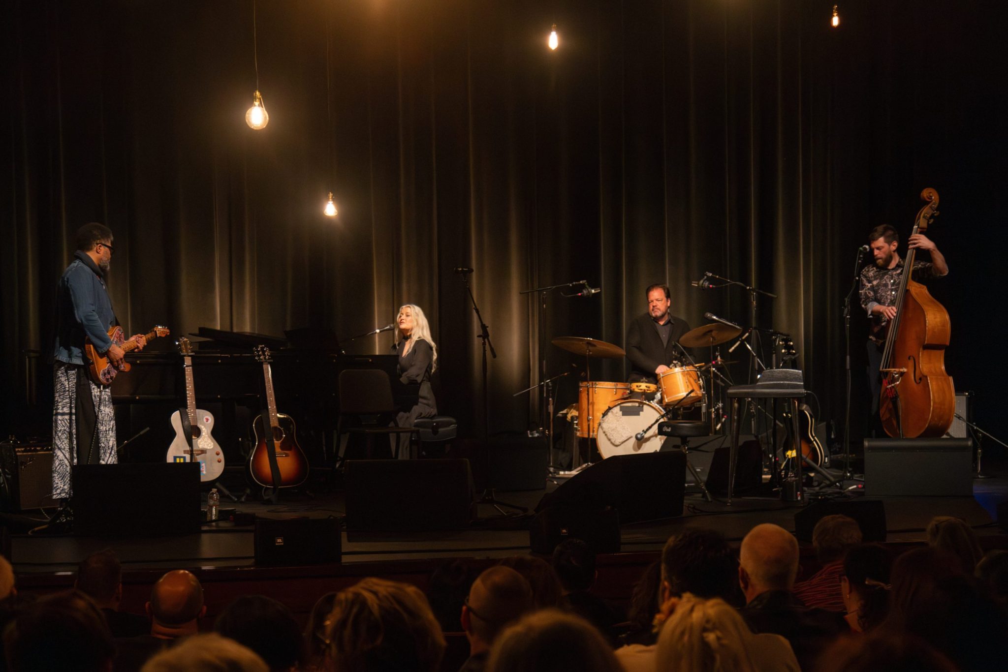 A band performs on stage, featuring a guitarist, a pianist/singer, a drummer, and an upright bass player.