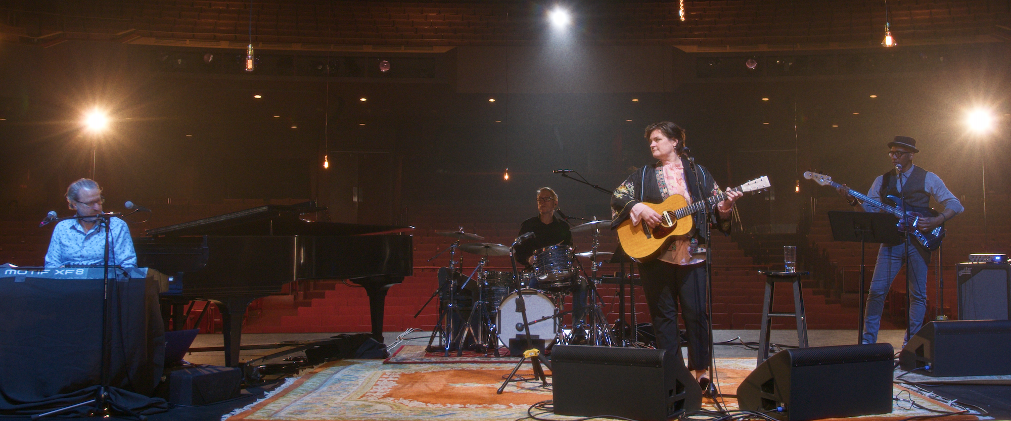 A band performs on a dimly lit stage with a pianist on the left, a drummer at the center, a guitarist singing in front, and a bassist on the right. The stage is adorned with rugs, and the venue has red seats and warm lighting.