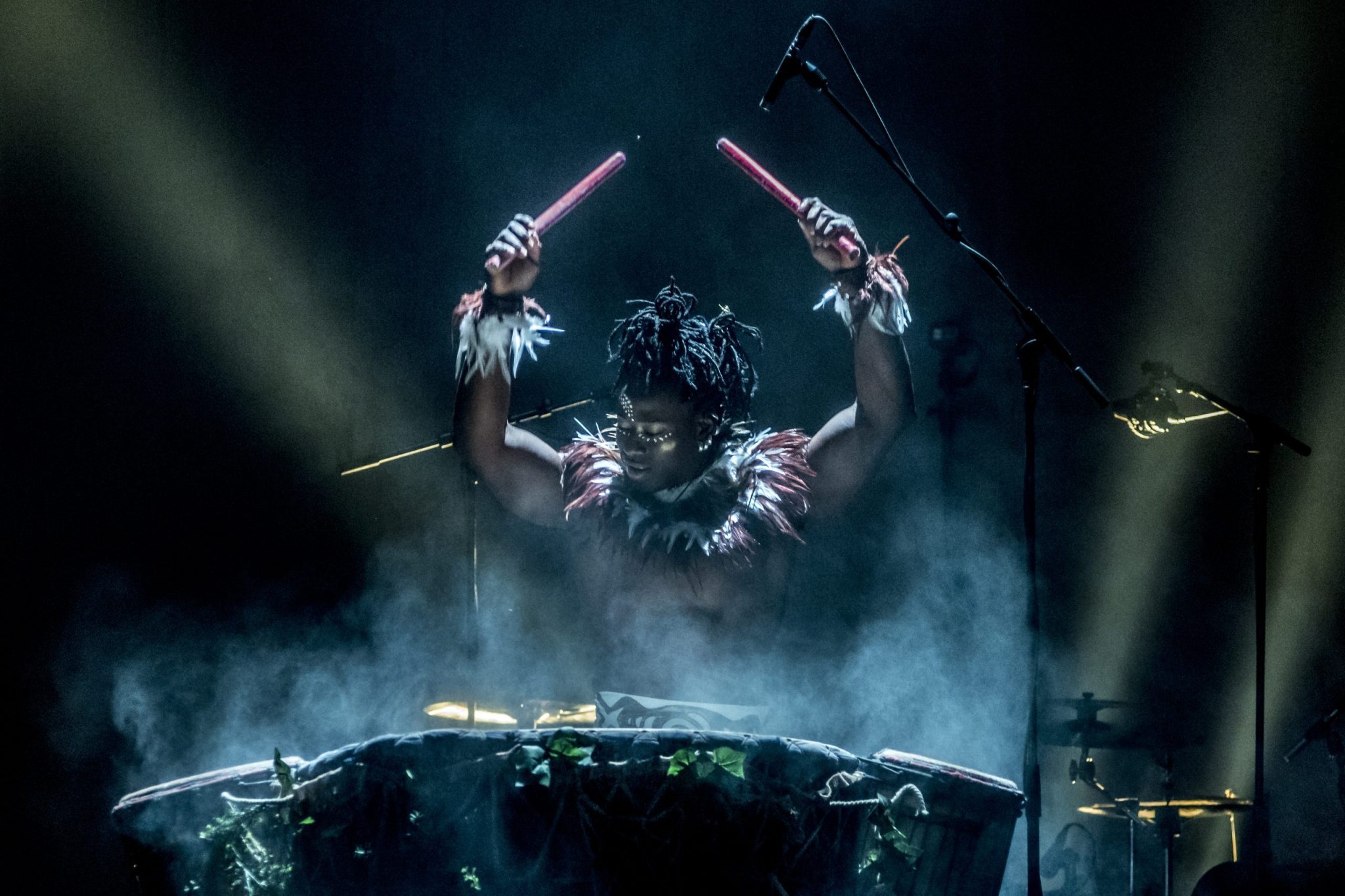 Cirque Kalabante performer wearing a headpiece and feathered attire raises drumsticks above their head while standing behind a large drum.