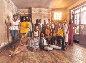 Kingdom Choir, a group of sixteen diverse individuals, dressed in stylish, elegant outfits in shades of white, yellow, and earth tones, pose together in a rustic room.