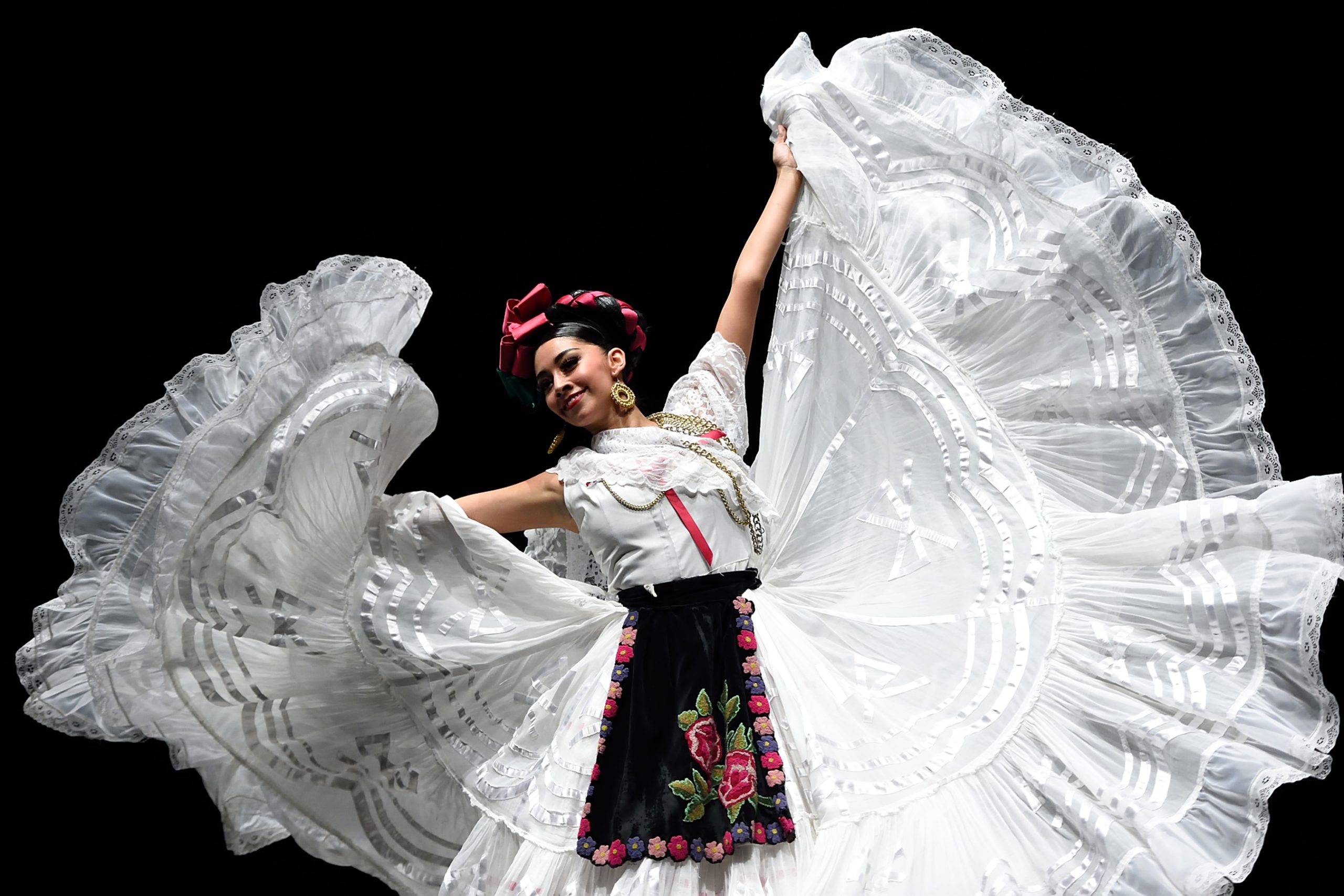 Ballet Folklorico dancer performs on stage in traditional Mexican attire, gracefully holding up a flowing white dress adorned with intricate embroidery.
