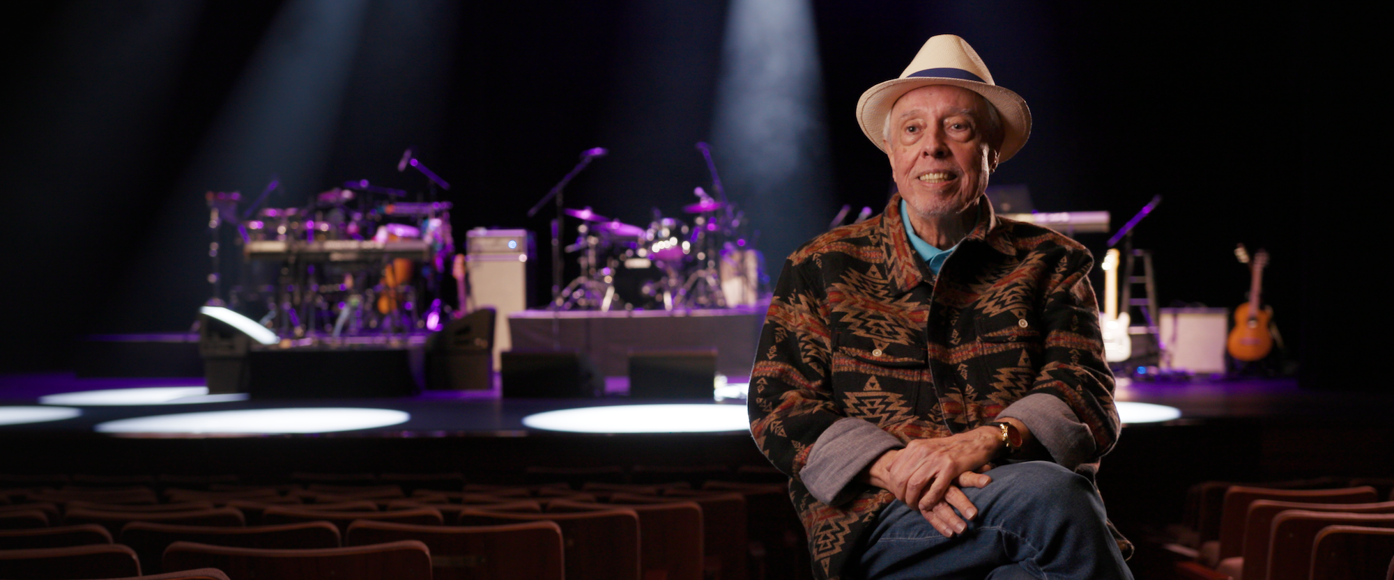 Sergio Mendes in a white hat and patterned jacket sits in a theater with a stage and musical instruments in the background. Stage lights create a dramatic ambiance.