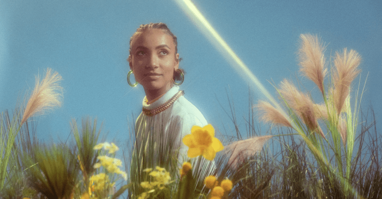 A woman with braided hair, wearing hoop earrings and a beaded necklace, looks off to the side while standing amidst tall grasses and yellow flowers. The bright blue sky and sunlight create a serene, dreamy atmosphere.