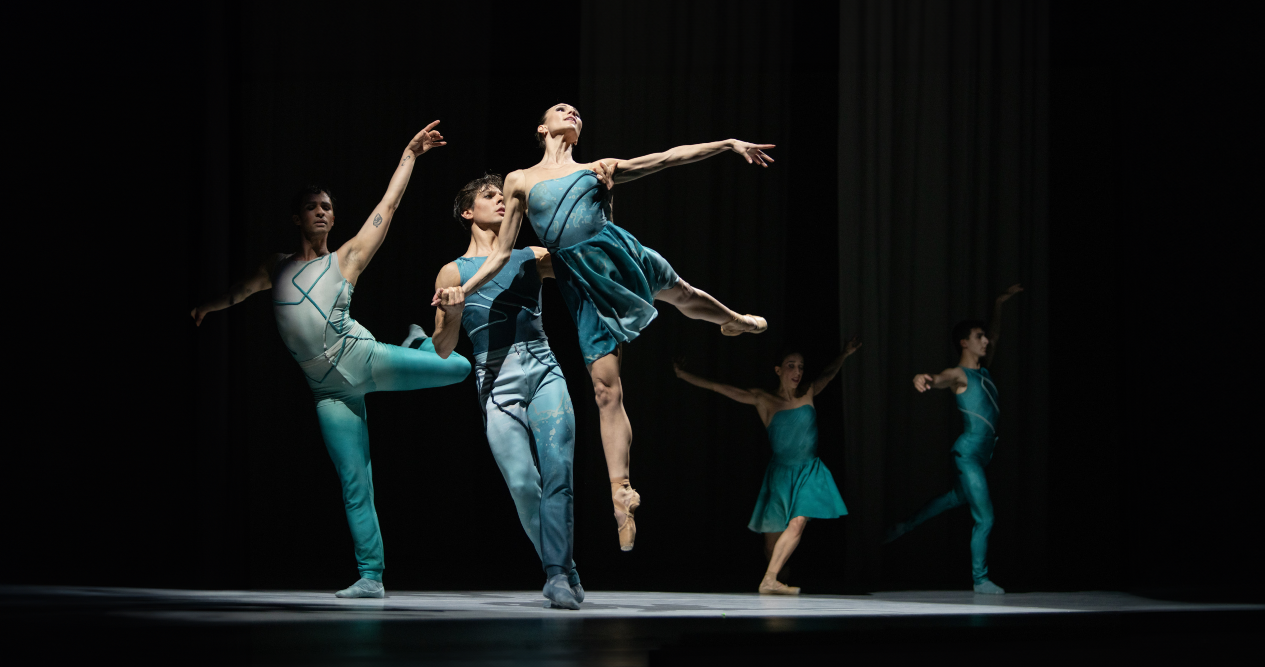 Five ballet dancers in teal perform; center duo lifted gracefully on dimly lit stage.