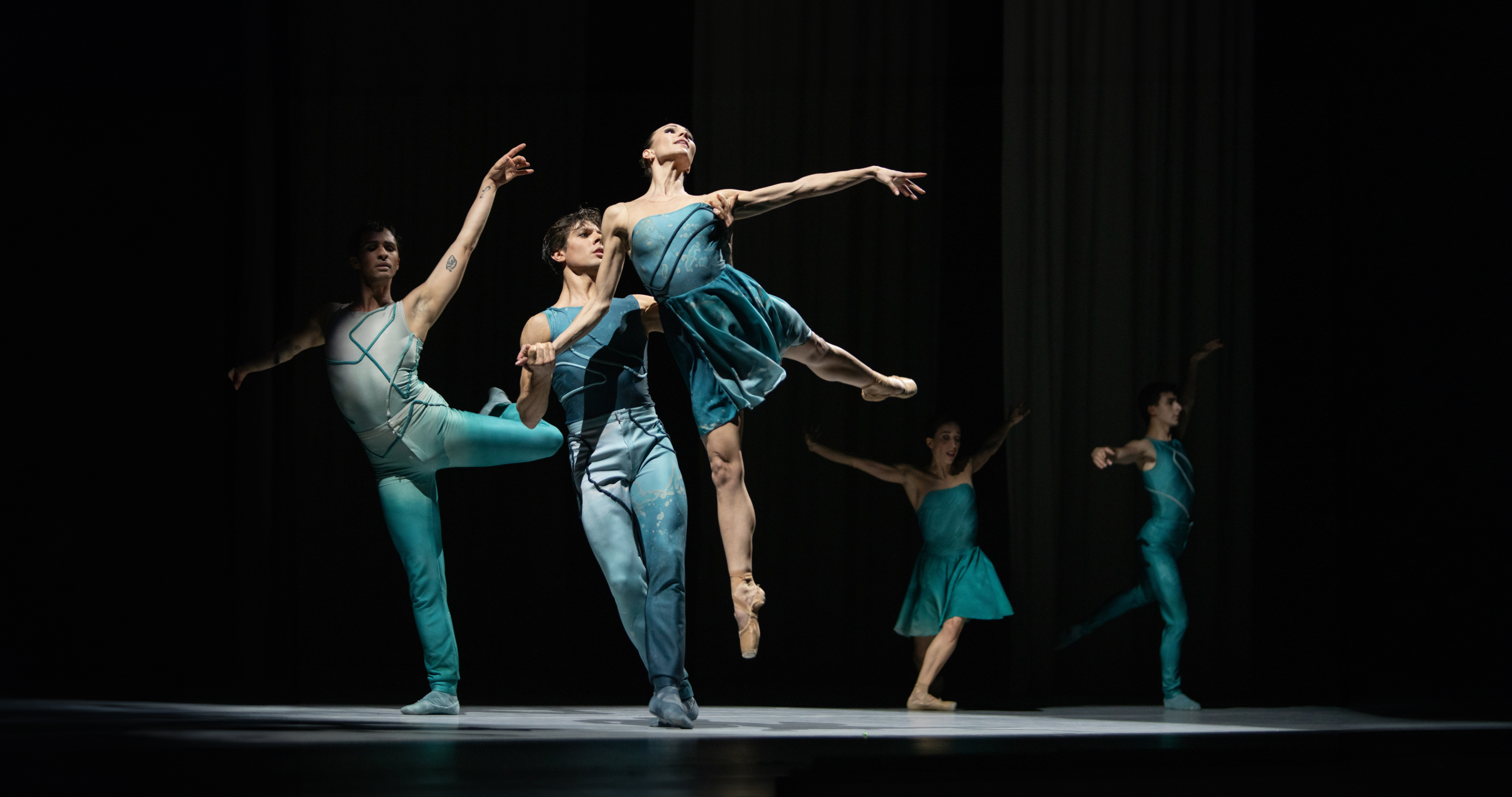 Five ballet dancers perform on stage in elegant poses. The central female dancer is lifted gracefully by a male partner. They all wear teal costumes, and the stage is dimly lit, highlighting their movements.