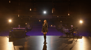 María José Llergo stands center stage in a dimly lit auditorium, singing into a microphone. She wears black attire and thigh-high boots. To her left, a person plays a keyboard, and to her right, another person plays a drum set. Hanging lights illuminate the stage area.