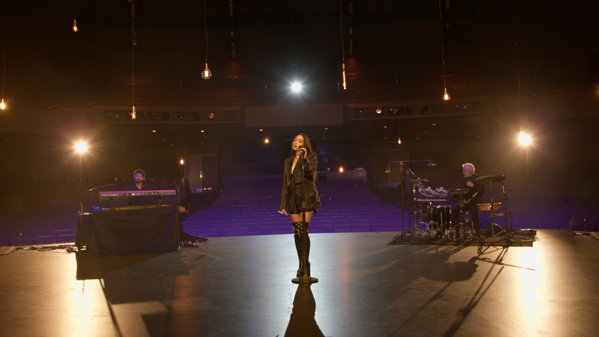 A singer stands on a dimly lit stage, illuminated by overhead lights. Musicians play on keyboards and drums in the background. The audience area is empty, and hanging light bulbs add to the ambiance.