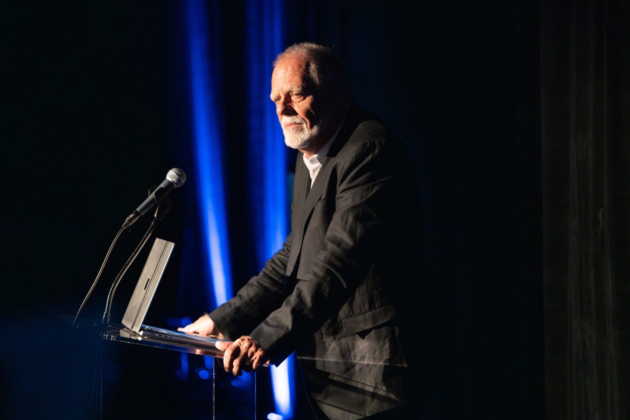 An older man with a beard stands in a dark suit behind a podium with a microphone and a laptop, speaking into a dimly lit room. Blue lights illuminate the background.
