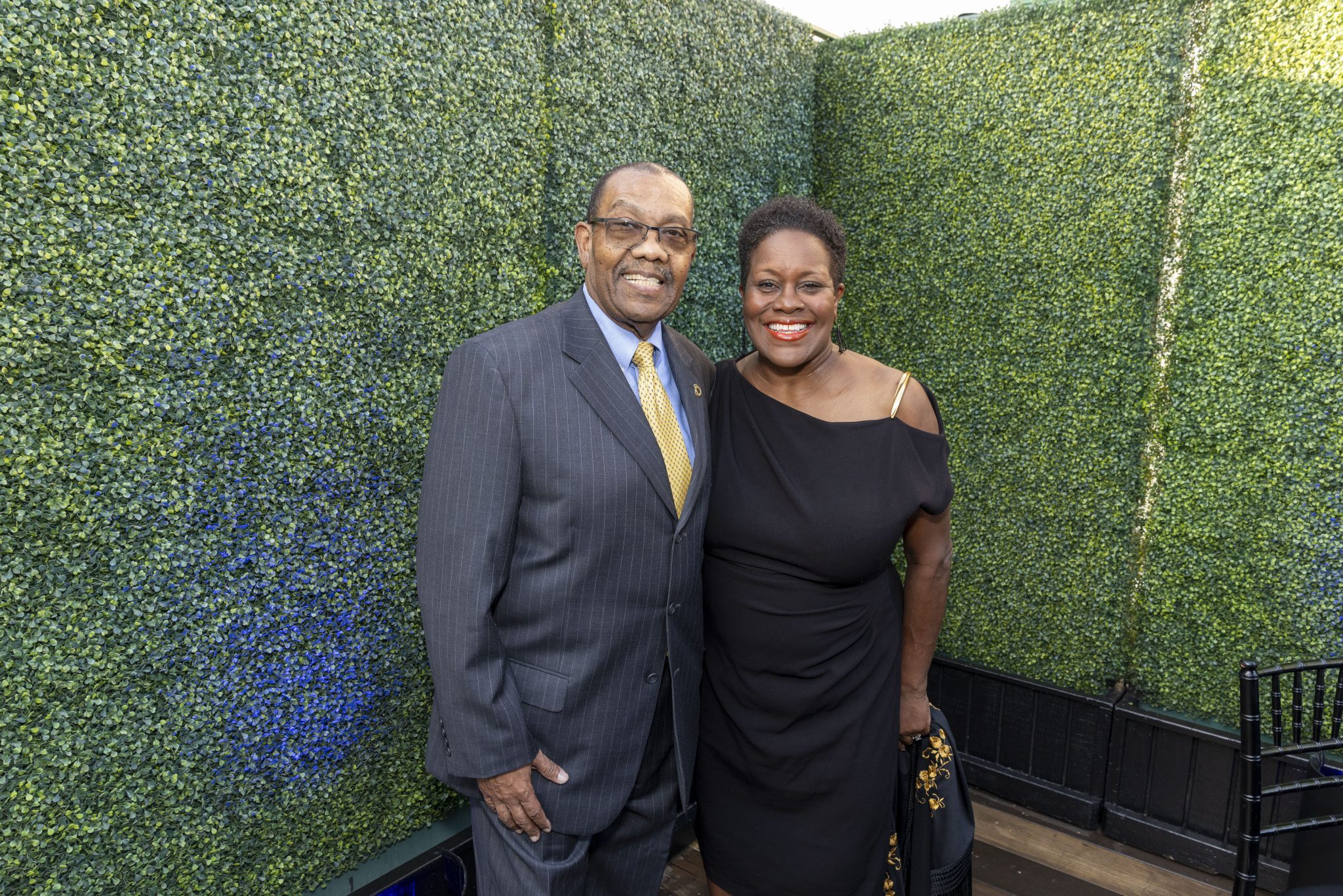 A man in a suit and a woman in a black dress smile while posing together in front of a leafy green wall outdoors.