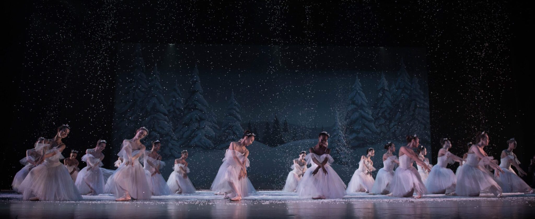 Ballet dancers in white costumes perform on stage, surrounded by a snowy backdrop and falling snow. The scene is reminiscent of a winter wonderland with dancers gracefully positioned in various poses.