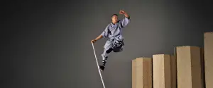 A monk in traditional attire leaps high, balancing on a staff with stacked wooden platforms behind.