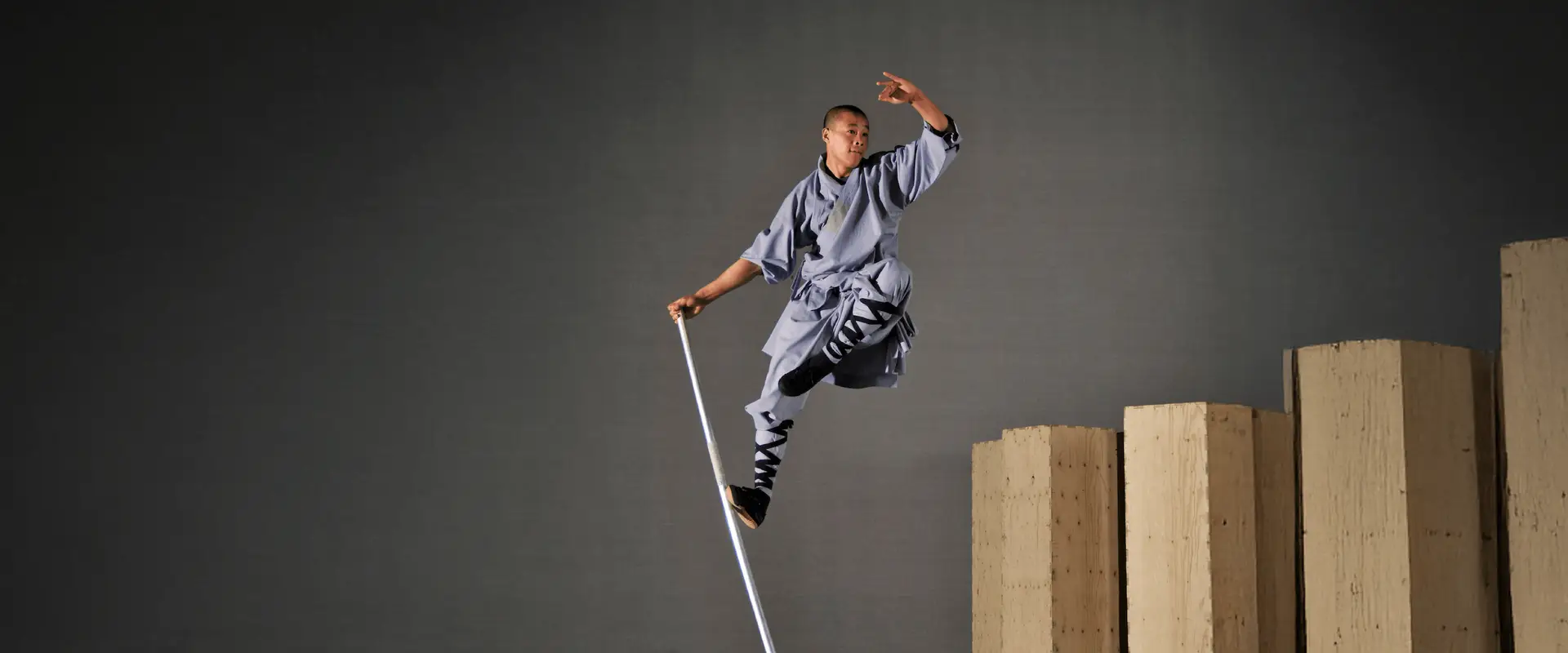A monk in traditional attire leaps high, balancing on a staff with stacked wooden platforms behind.