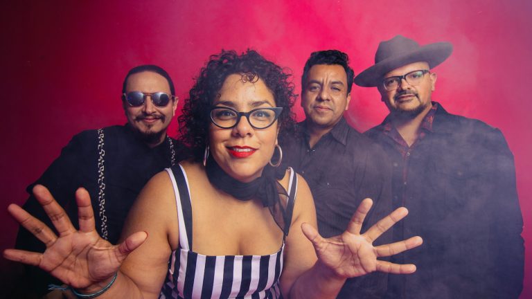La Santa Cecilia in black outfits pose against a smoky pink background, with the woman in front.