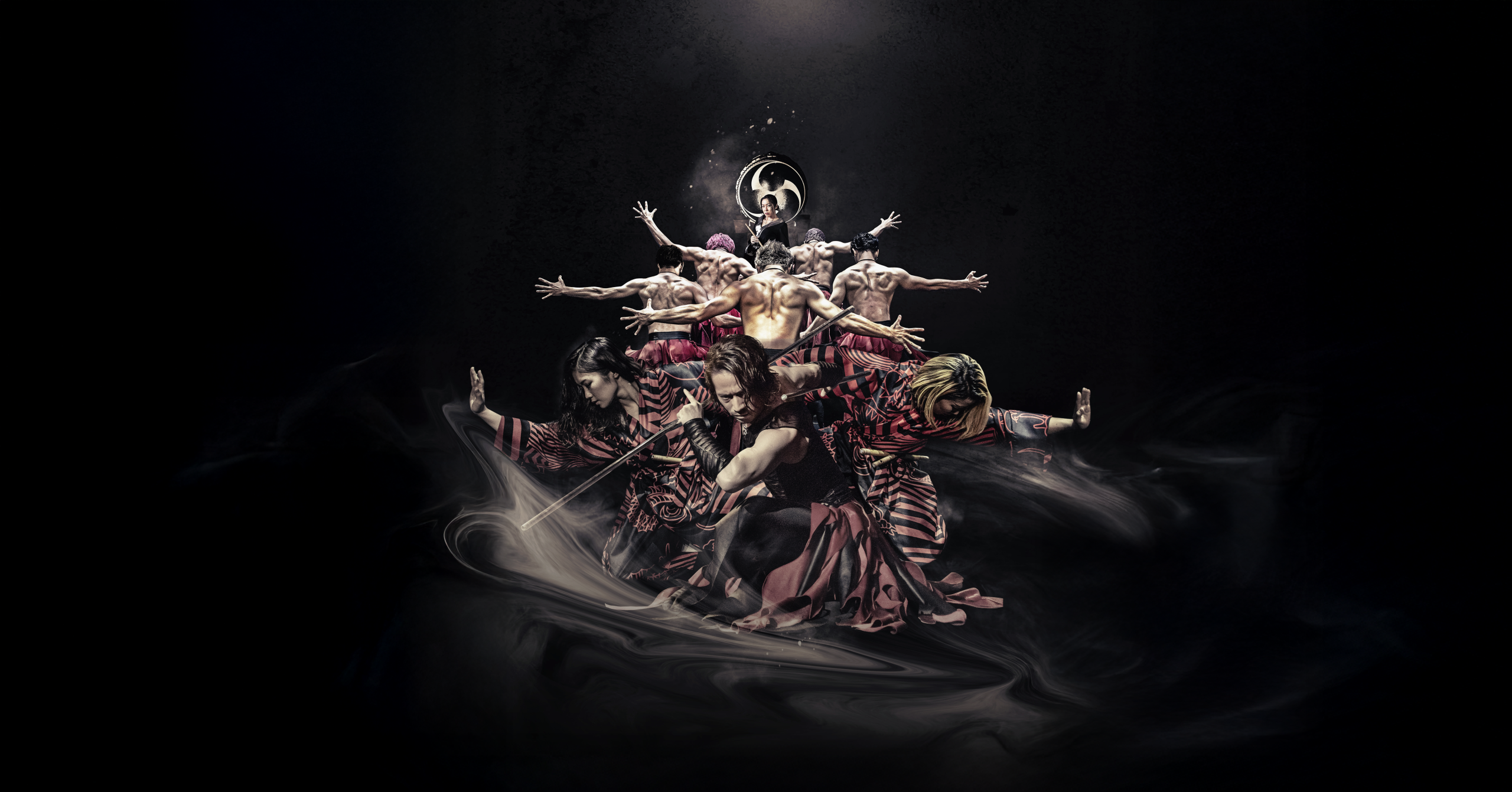 A group of performers in dramatic poses, some shirtless and others in patterned outfits, arranged in a layered formation