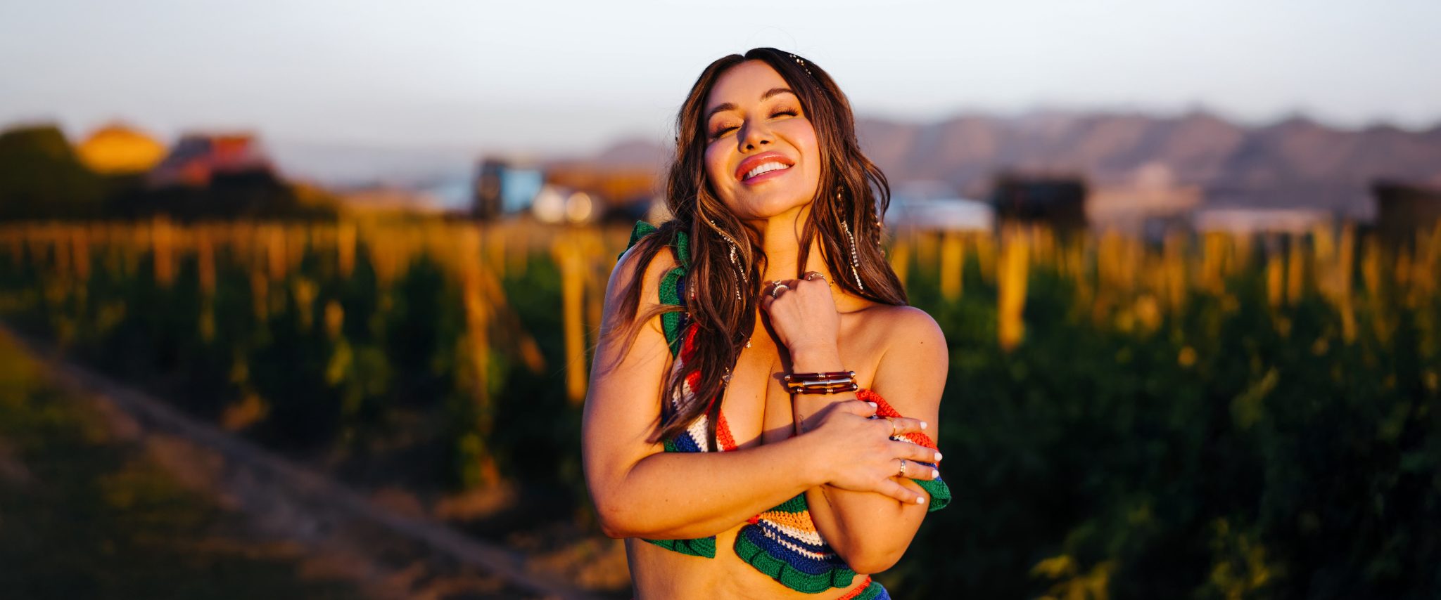 Chiquis standing in a vineyard at sunset, smiling with her eyes closed and arms crossed over her chest.