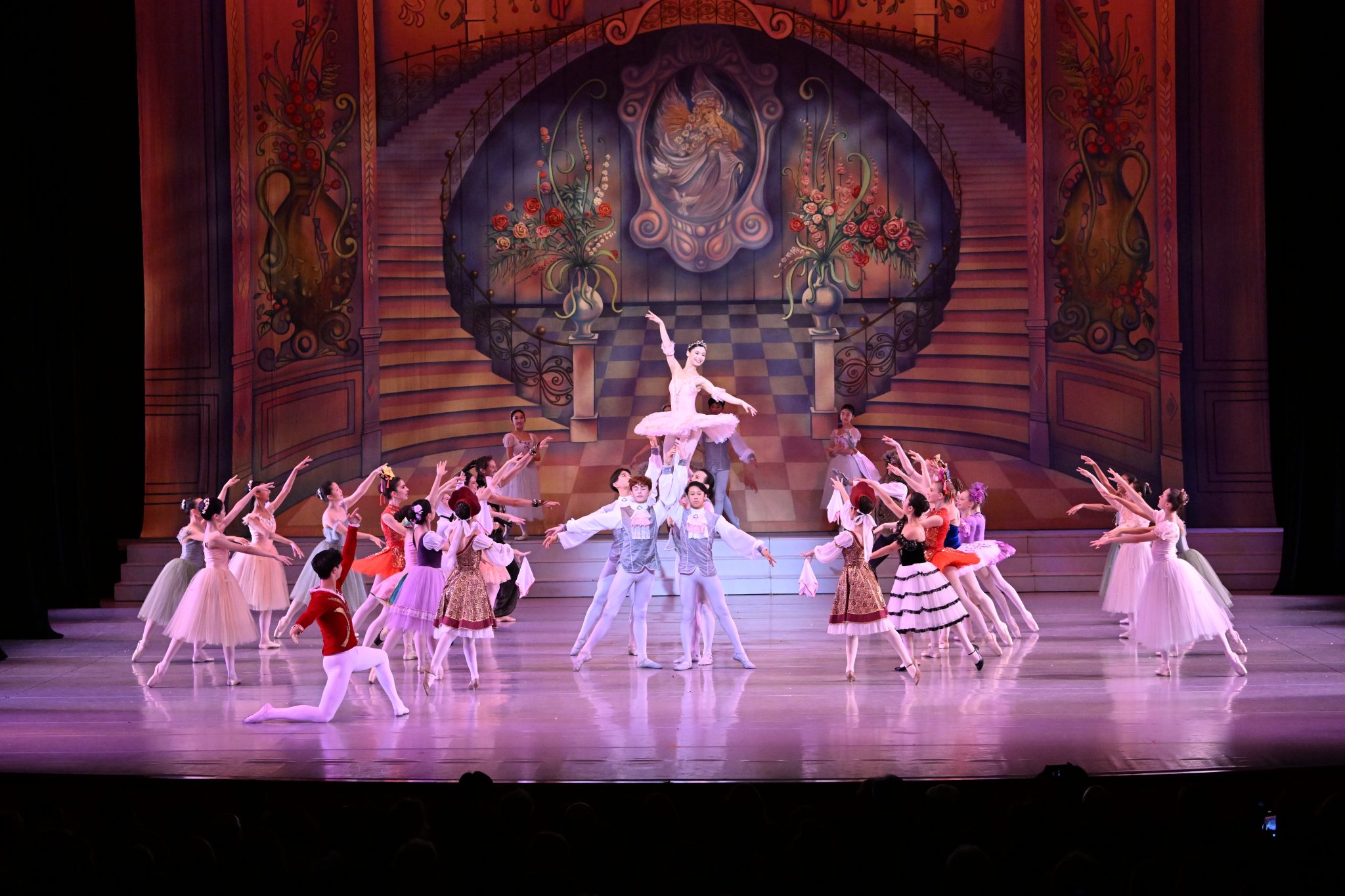 Ballet dancers perform on stage in elaborate costumes with a painted backdrop featuring stairs and floral decorations.