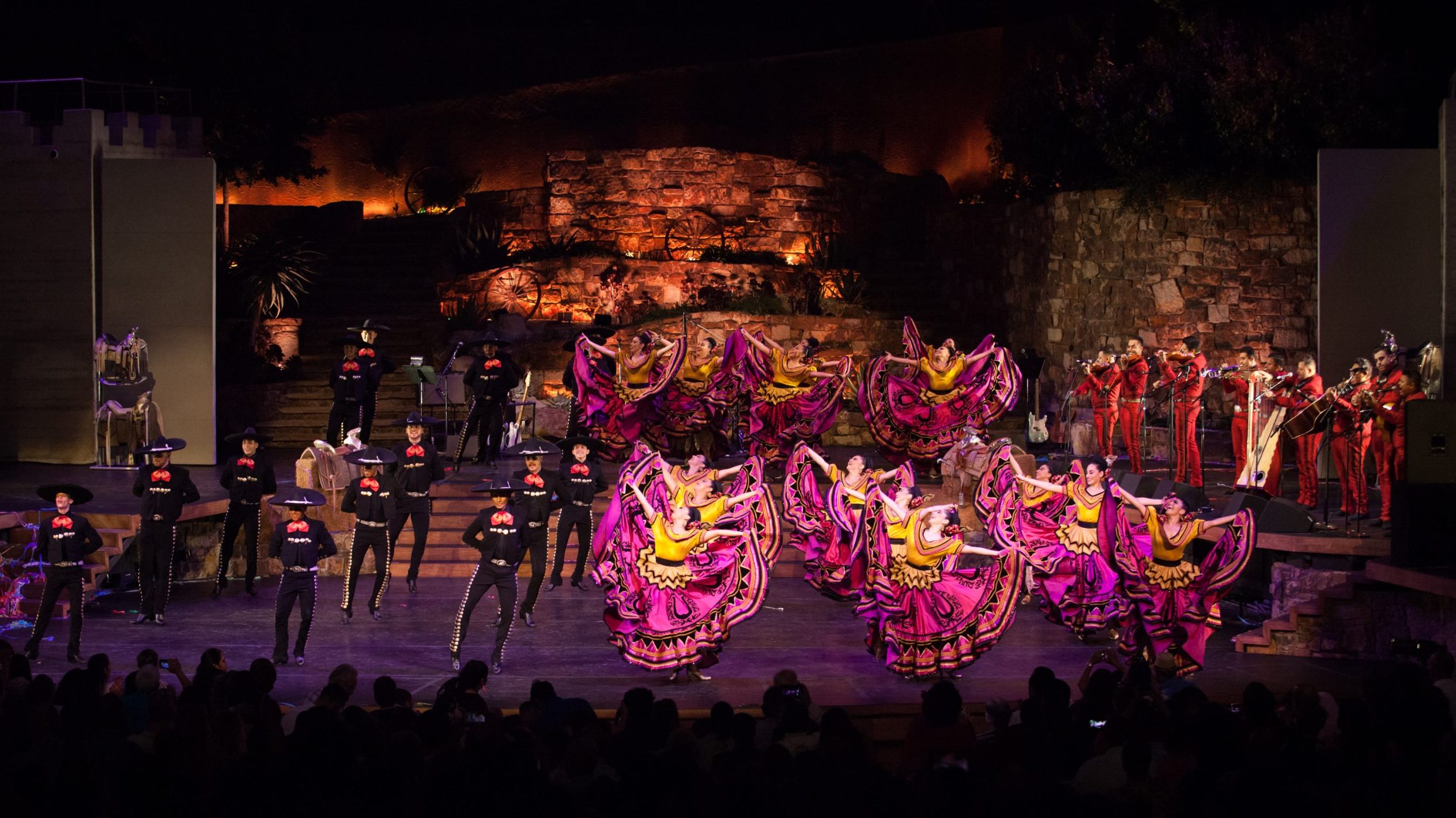 A group of folklorico dancers in colorful traditional dresses perform on stage with a mariachi band in the background, under stage lights.