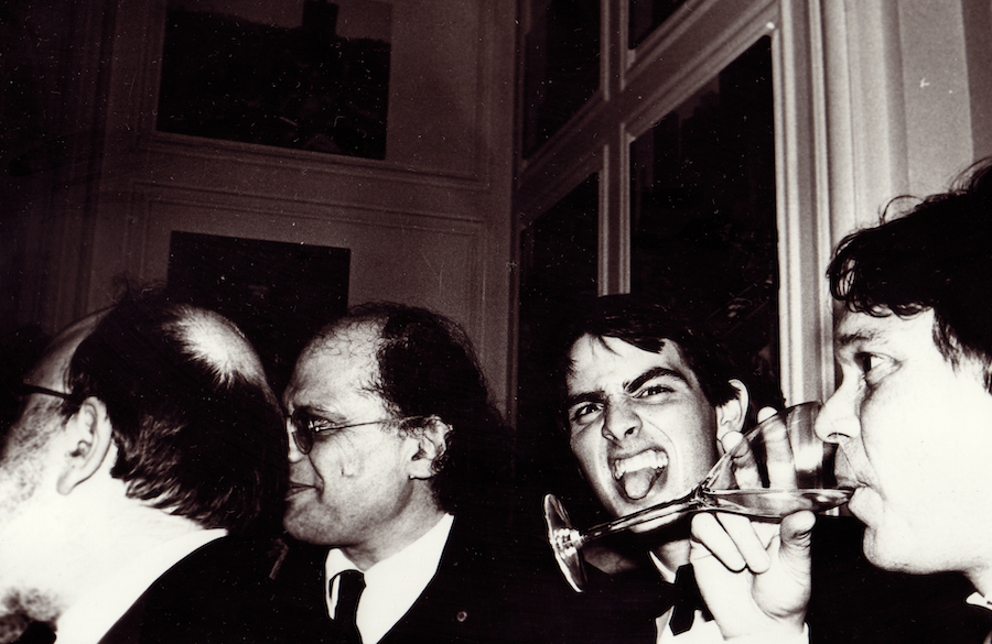 Four men in formal attire stand closely together; one man in the middle is sticking out his tongue while another drinks from a wine glass.