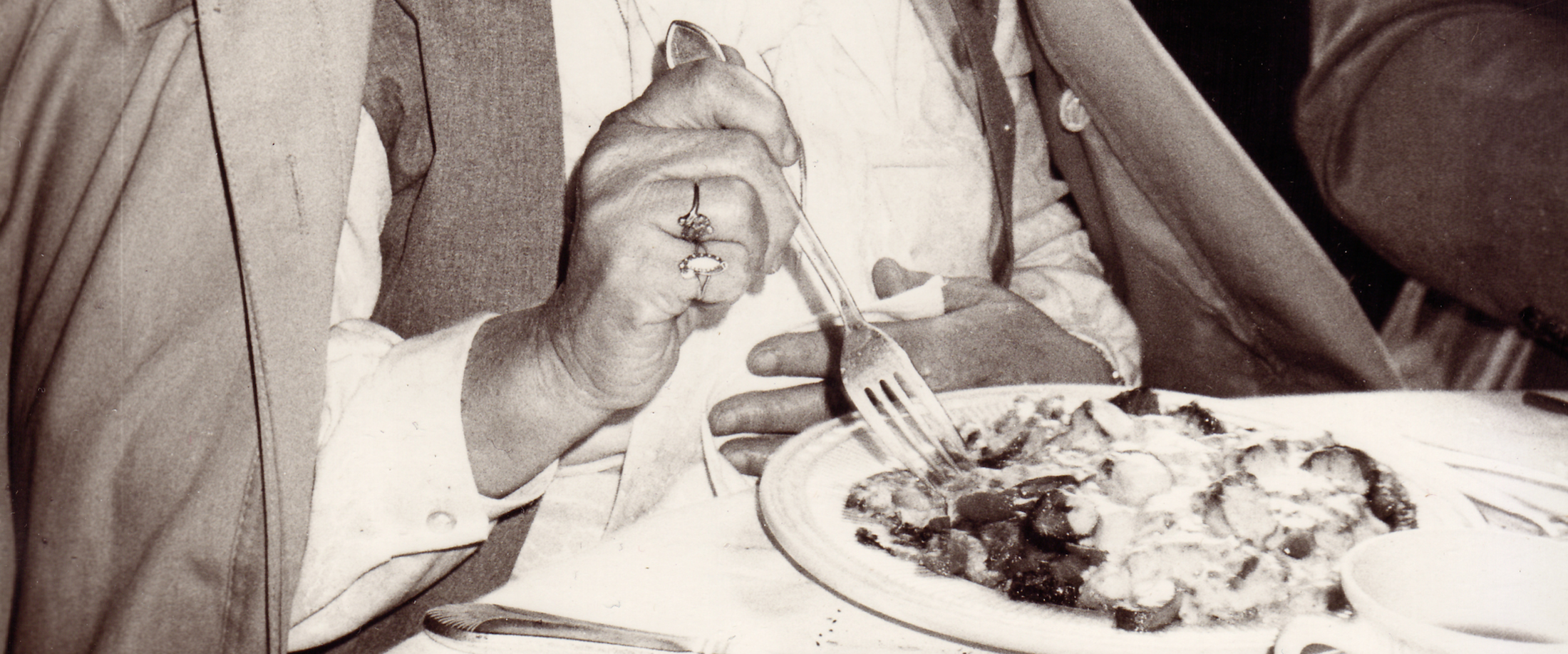 A person wearing a ring uses a fork to eat food from a plate at a table, dressed in a suit and collared shirt.
