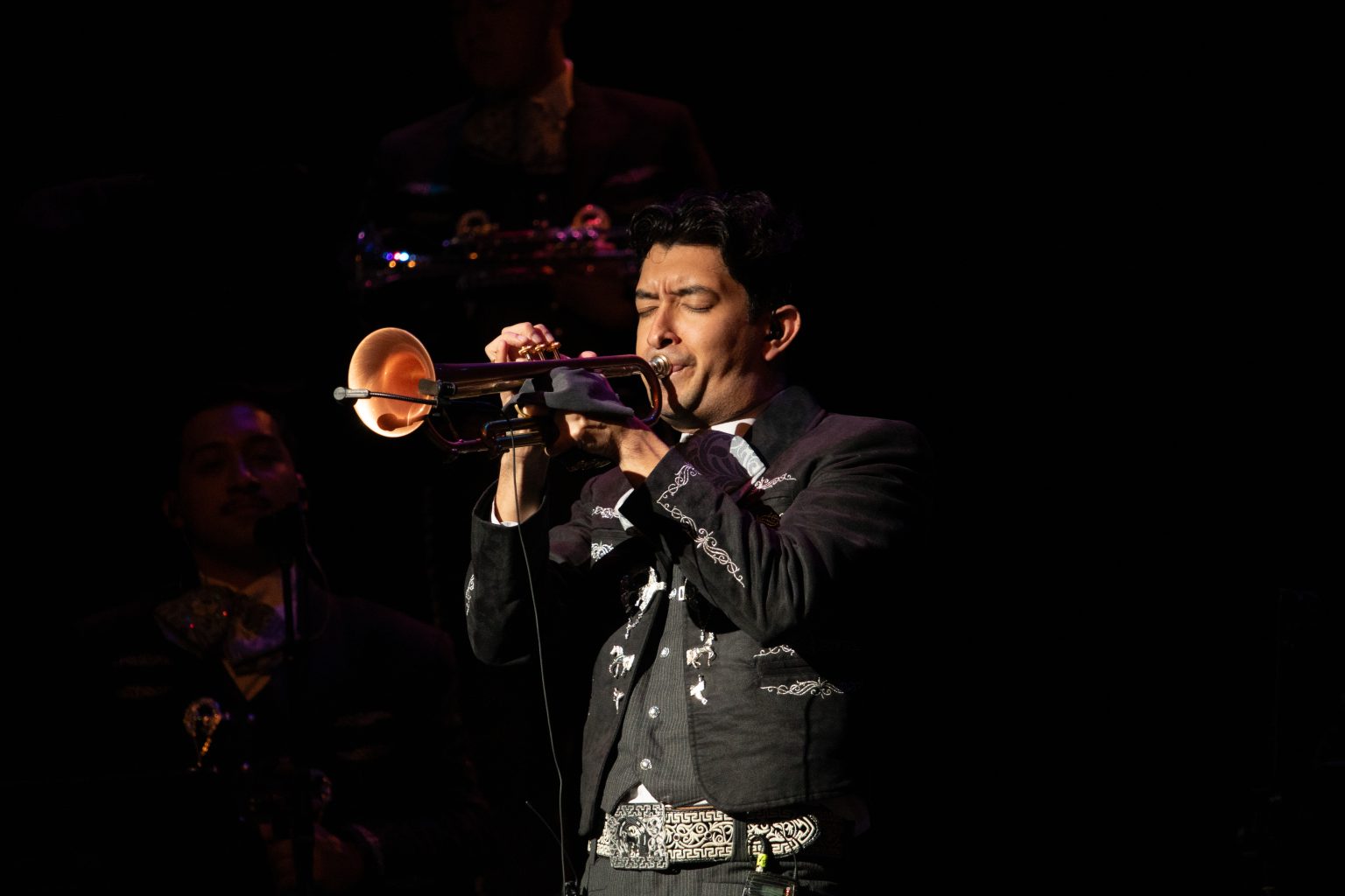 A mariachi musician in a traditional charro suit plays the trumpet on a dimly lit stage.