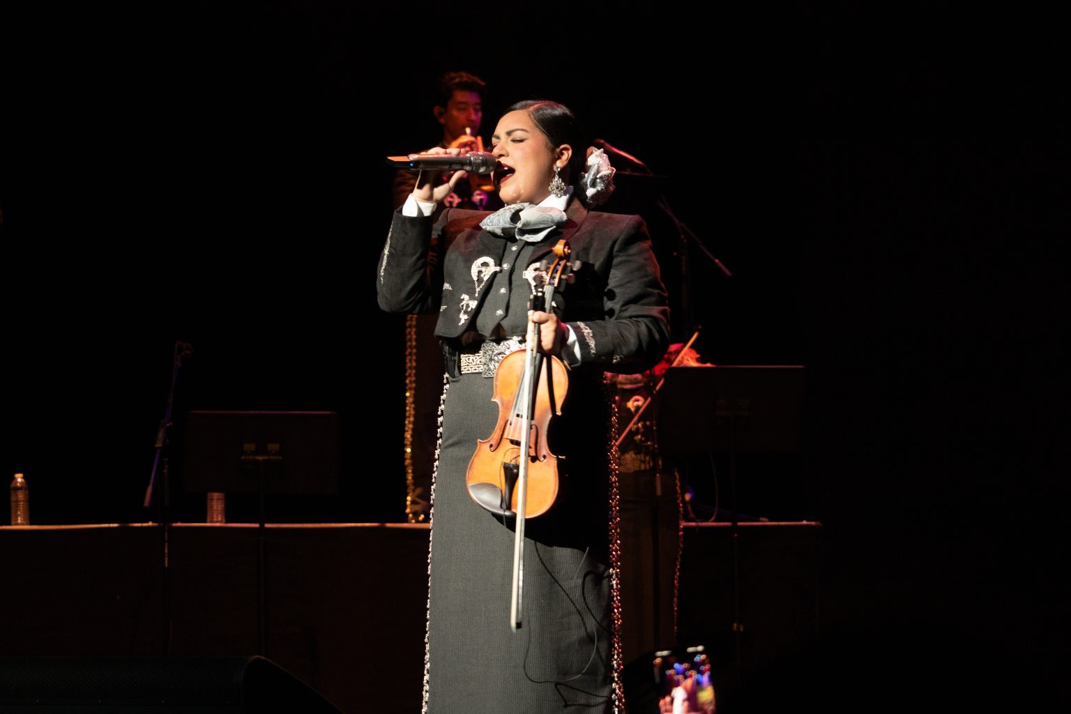 A mariachi musician in traditional attire sings into a microphone on stage while holding a violin; other musicians are visible in the background.