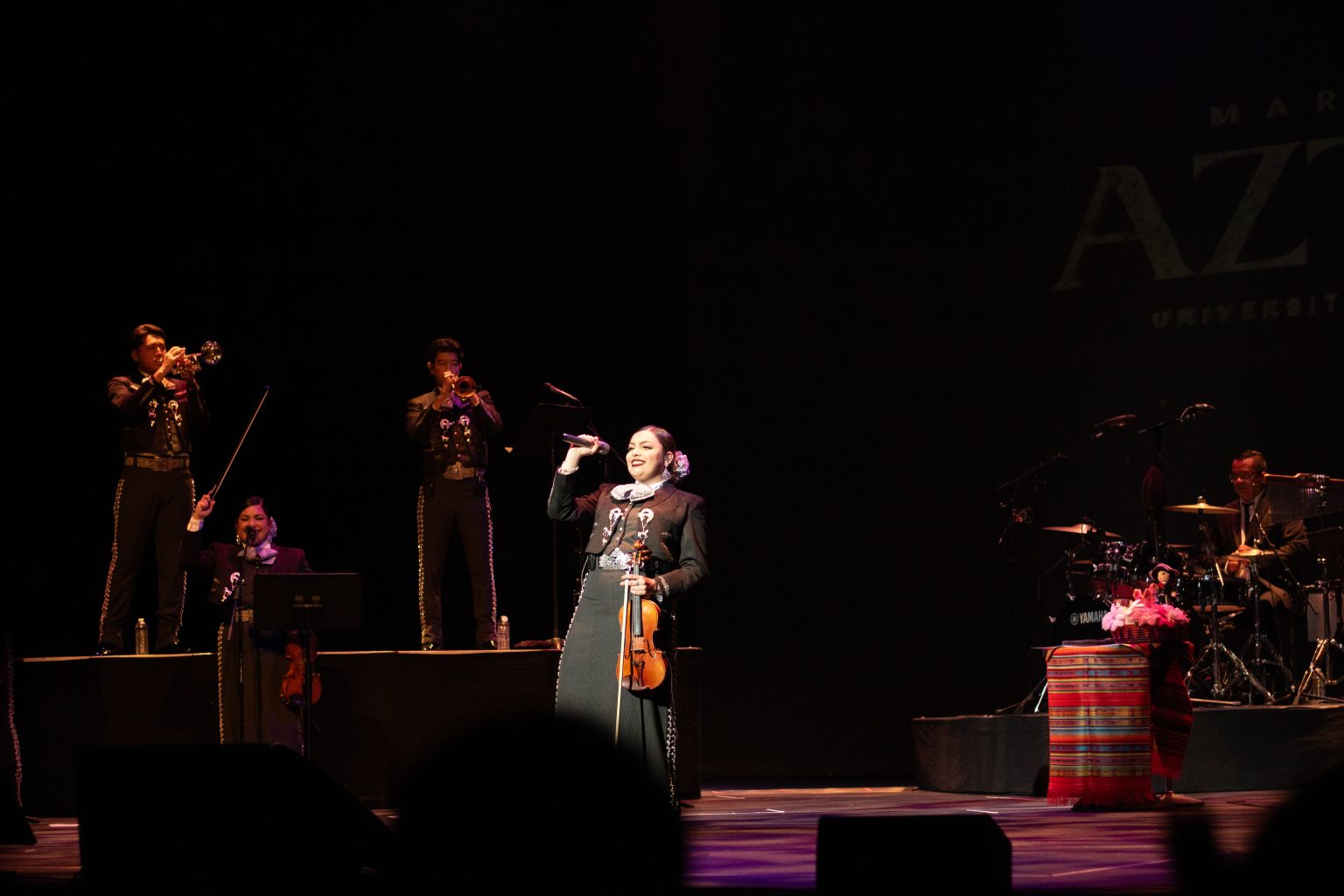 A mariachi band performs on a dimly lit stage, with a female lead singer holding a microphone and violin at center stage.