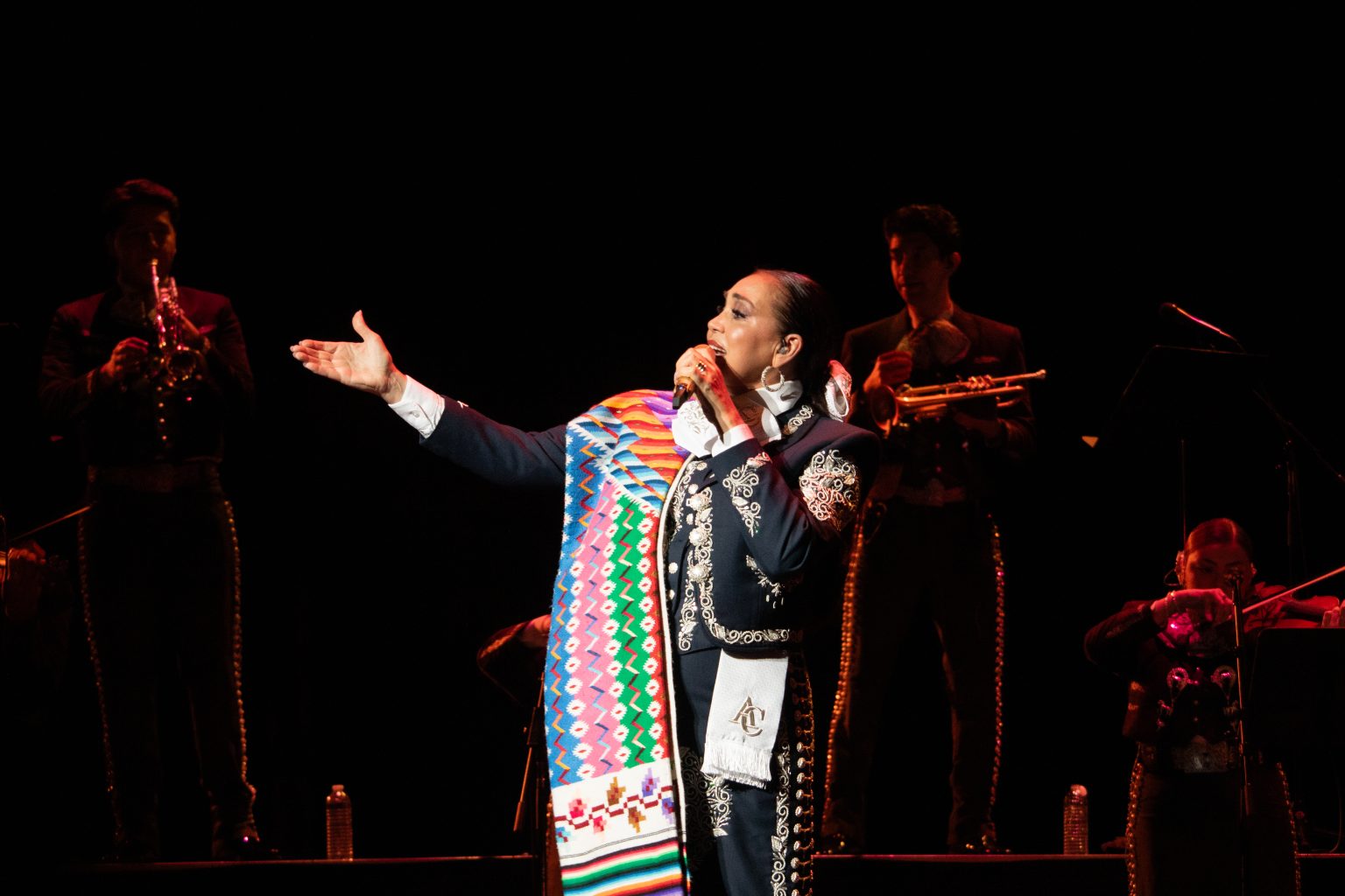 Aida Cuevas in a traditional outfit and colorful sash performs on stage, accompanied by musicians playing brass instruments in the background.
