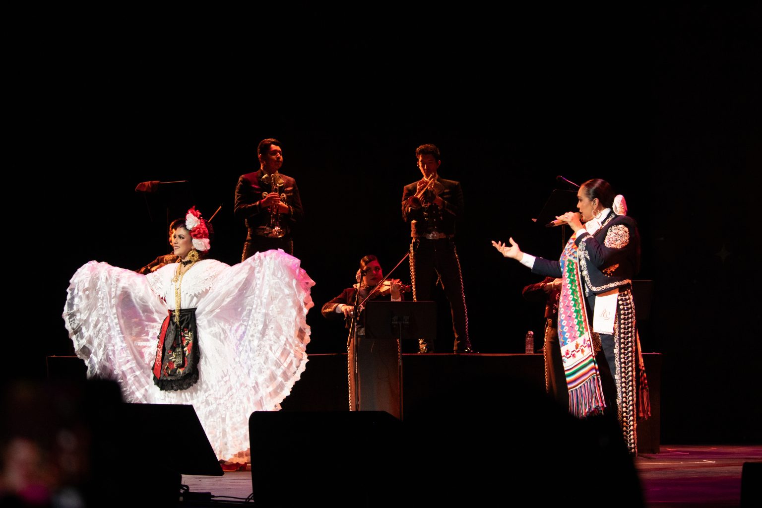 Two women in traditional Mexican dresses perform on stage with three musicians in black suits playing instruments behind them.