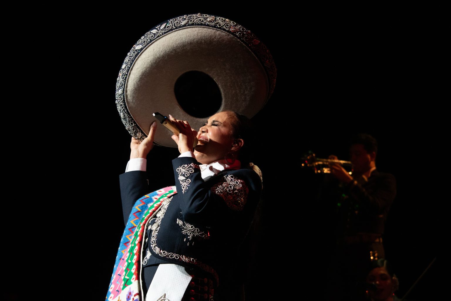 Aida Cuevas in mariachi outfit performs passionately on stage, holding a sombrero above their head, with a trumpet player in the background.