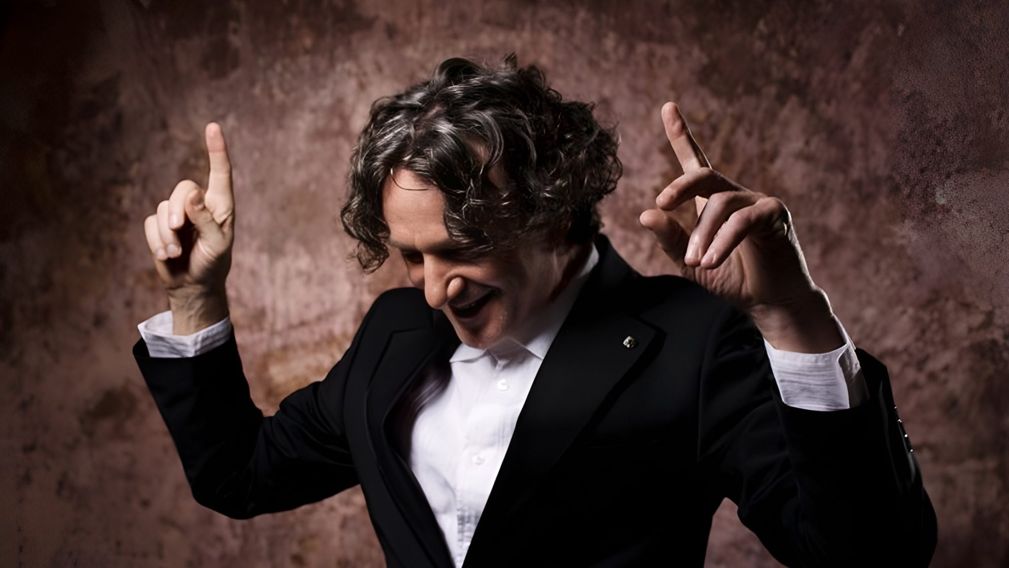 Goran Bregović in a black suit with a white shirt stands against a textured brown background, smiling and raising both hands with index fingers pointing upward.