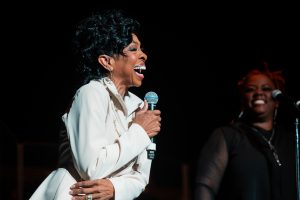 Gladys Knight in in a white outfit holds a microphone and sings on stage, smiling, with another person in black standing and smiling in the background.