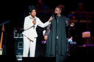 Gladys Knight and backup singer stand and sing on stage, one in a white suit holding a microphone and the other in a long black dress, with musicians and instruments visible in the background.