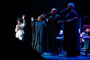 Gladys Knight and backup singers stand on stage under spotlights, each with a microphone, performing together; one singer is in white while the others wear black.