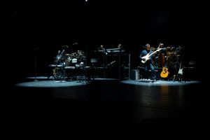 A band performs on a dark stage with various instruments, including keyboards, drums, electric bass, and guitars, illuminated by spotlights.
