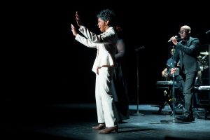 A woman in a white suit performs on stage with a microphone, accompanied by a band and backup singer, under stage lighting.