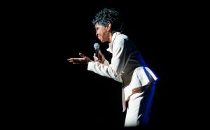 Gladys Knight in a white suit stands on stage holding a microphone, gesturing with one hand, against a dark background.