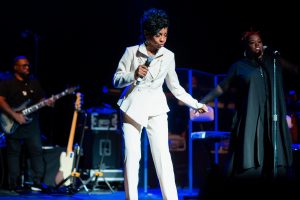 Gladys Knight in a white suit performs on stage holding a microphone, with a guitarist and backup singer in the background.