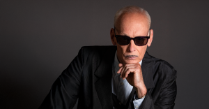 John Waters wearing dark sunglasses, a black suit, and a light shirt, poses with their hand on their chin against a plain dark background.