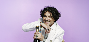 A man with curly hair in a white suit smiles while resting his arm on a bottle against a purple background.