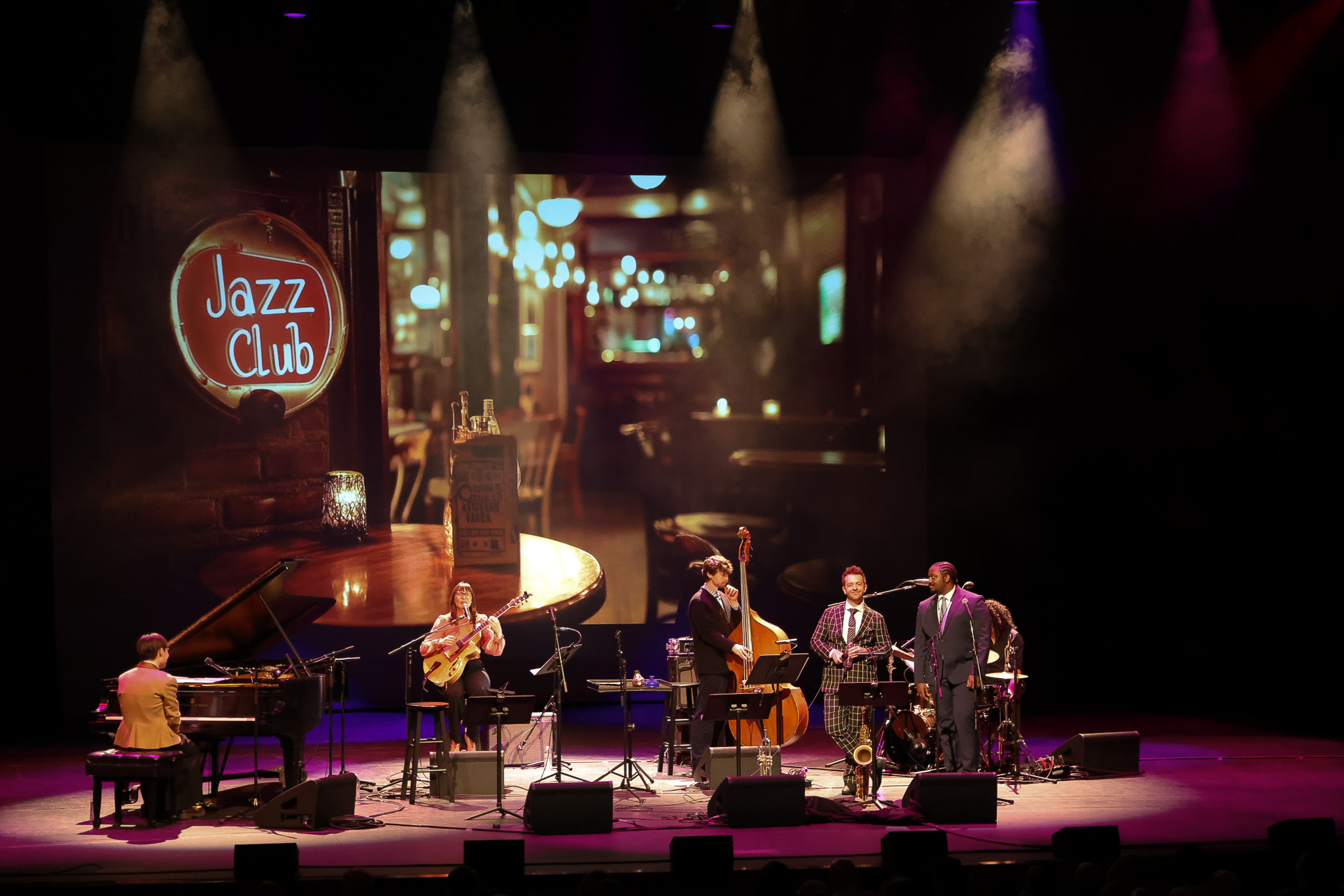 A jazz band performs on stage under purple lighting with a "Jazz Club" sign and bar scene projected in the background.