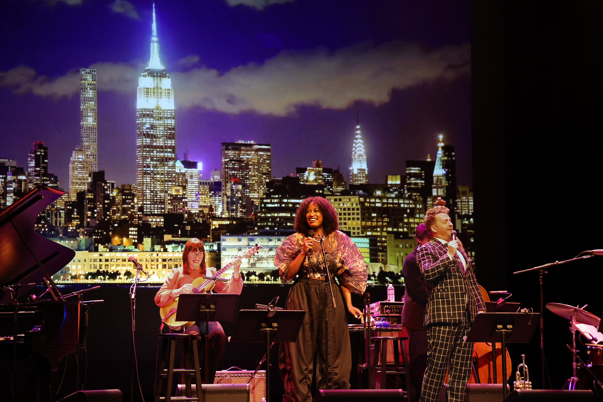 Musicians perform on stage in front of a backdrop featuring a nighttime city skyline with the Empire State Building and Chrysler Building visible.