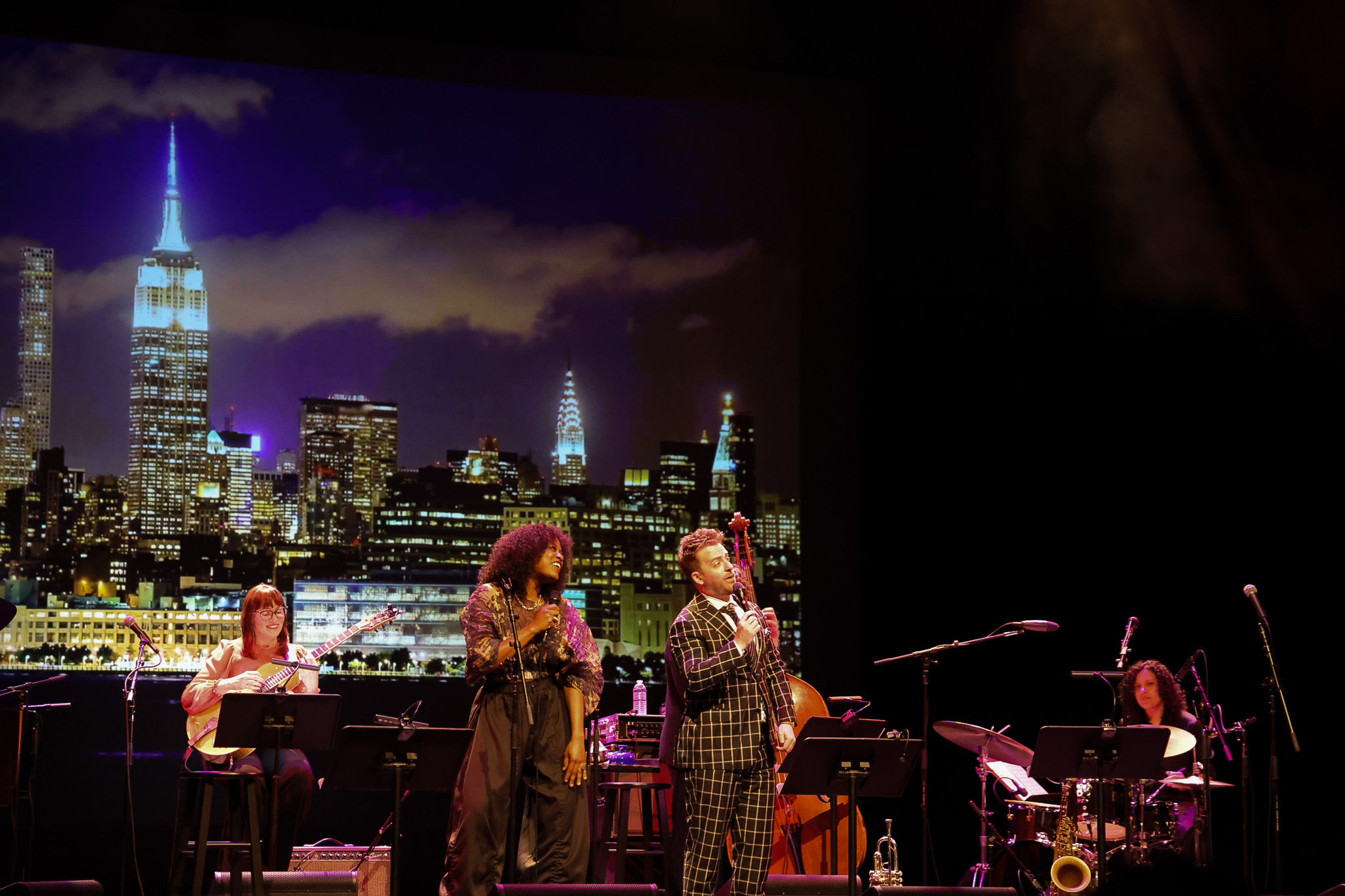 Musicians performing on stage with a New York City skyline backdrop, including vocalists at center and instrumentalists seated on either side.